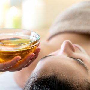 Macro close up of hand holding glass bowl with aromatic oil next to woman’s head in spa.