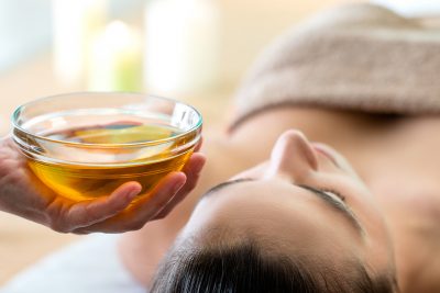 Macro close up of hand holding glass bowl with aromatic oil next to woman’s head in spa.